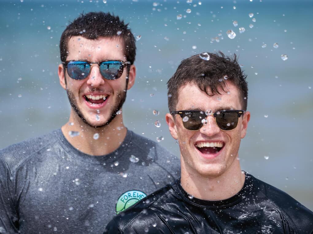 Two men at the beach wearing sunglasses and t-shirts with water droplets sprayed across their faces